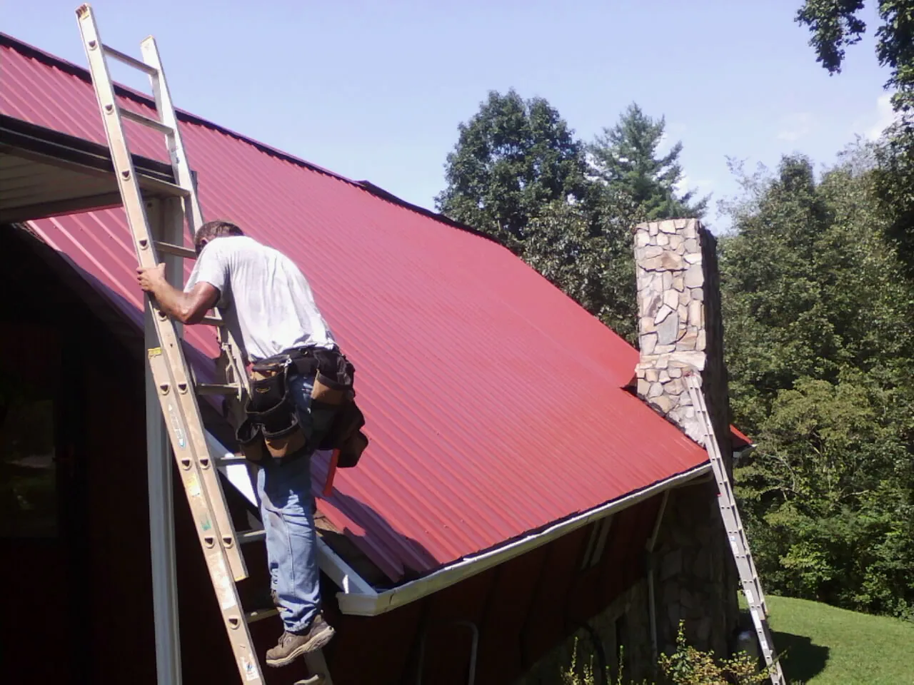 Roofer on a ladder beside a red metal roof and stone chimney — Soffit & Fascia Repair by Roofing Brothers in Prospect Heights